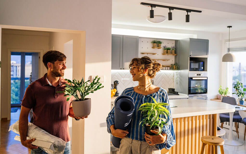 A happy couple smiles at each other as they carry house plants and other items into the home