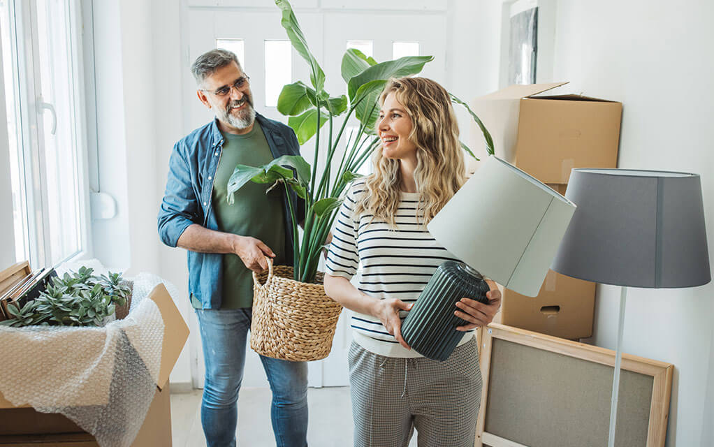 An older couple carrying their stuff into their new home smile as the woman leading carries a lamp and the man carries a potted plant as open boxes and other items line the entryway