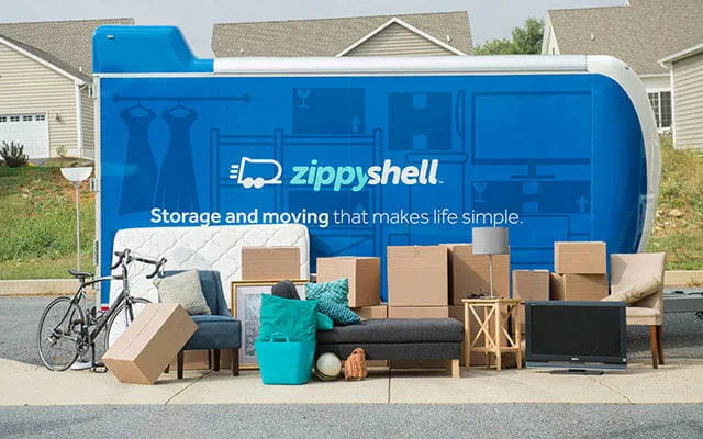 A Zippy Shell portable storage container sits in front of a suburban neighborhood's home on a sunny day with a homeowners furniture and belongings staged in front of it from a side view perspective