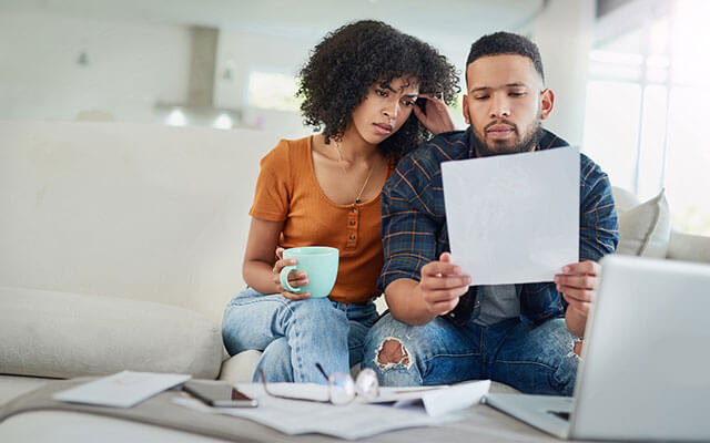 A couple sits on a couch in their home examining a piece of paper looking concerned with a laptop, cell phone, and other papers sitting in front of them