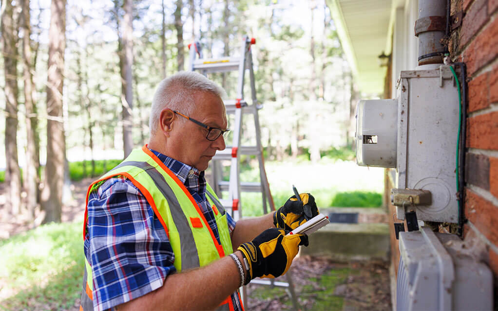 An older man in a reflective vest holds a clipboard and makes notes while inspecting the outside of a home and its power input site