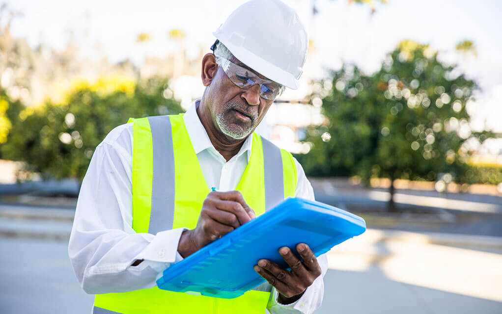 An older man wearing a hard hat and reflective vest makes notes on the clipboard he is holding
