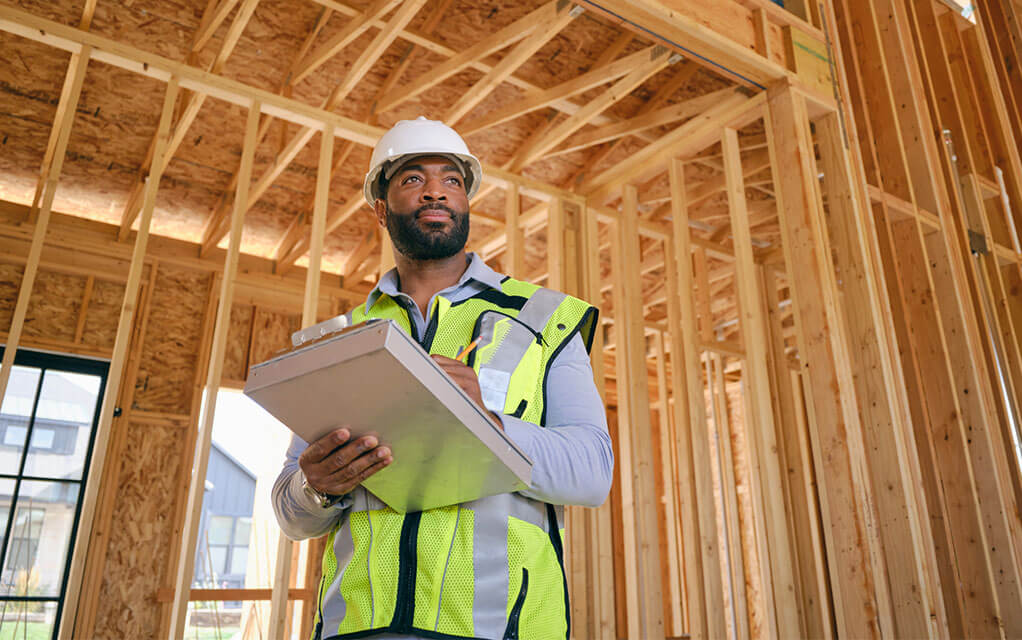 A home inspector wearing a white hard hat and a reflective yellow jacket holds a clipboard and makes notes in an unfinished home
