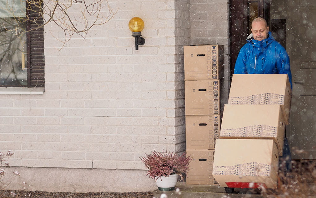 A professional laborer wearing a waterproof jacket moves boxes out of a house on a moving dolly as snow falls outside