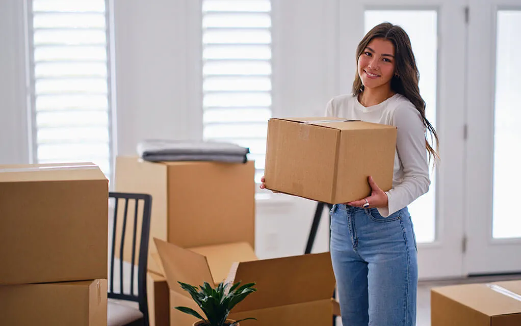 A cheerful woman holds a packed box in her arms in her home's living room as she is surrounded by other packed and unpacked boxes on the floor