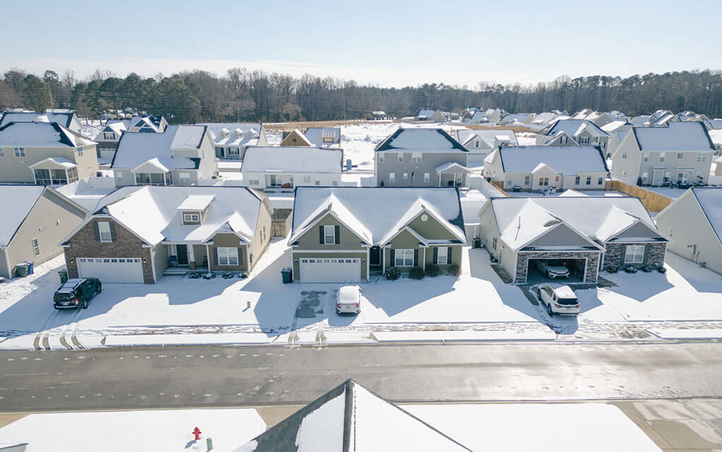 Aerial view of an American neighborhood that is covered in snow, including houses and vehicles, as the sun shines down on a clear day