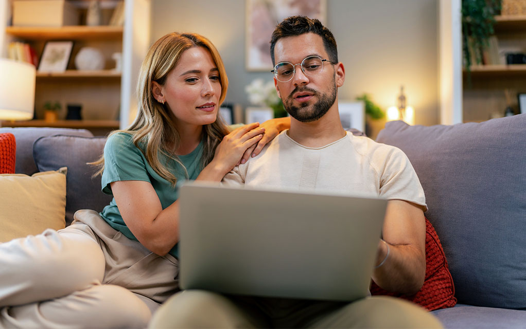A young couple sits on their couch looking at a laptop for something important in their home's living room
