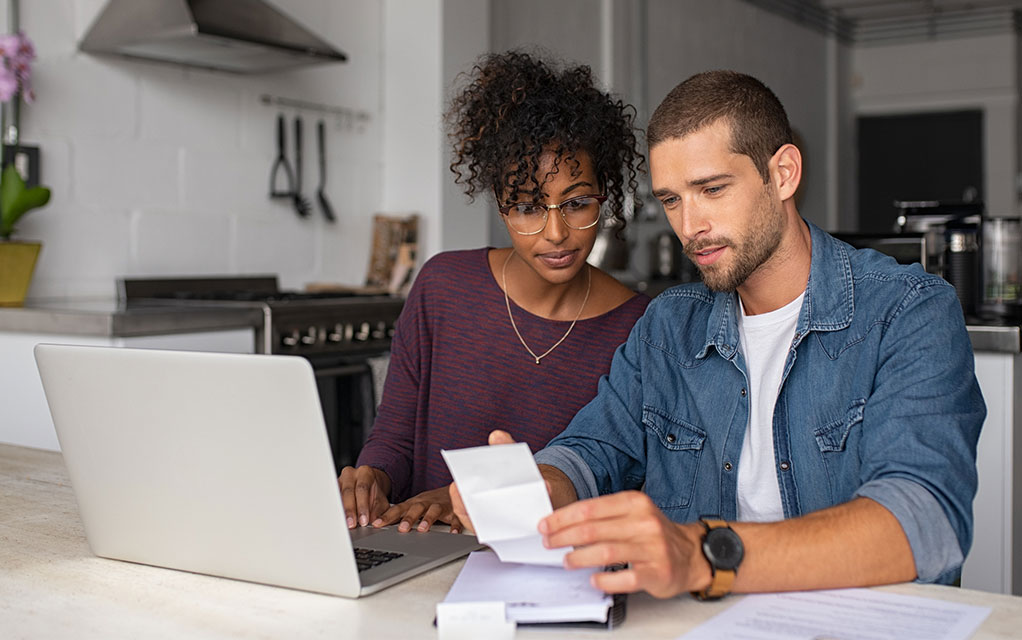 A couple looks at receipts and a budget ledger with an open laptop in the home's kitchen at a table