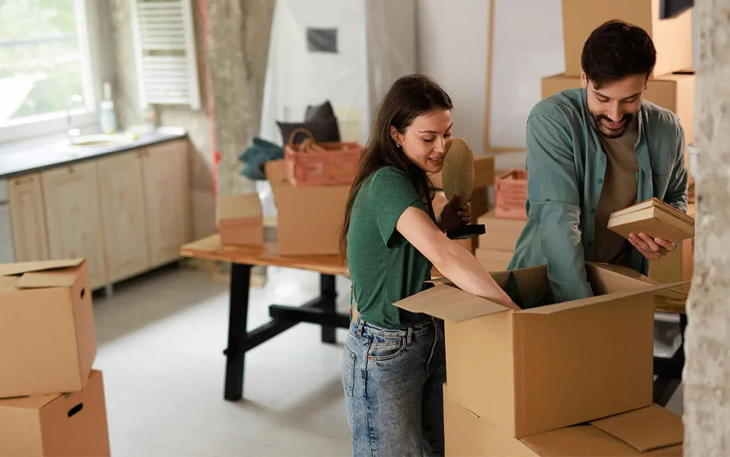 A couple is removing items out of a moving box in their new home, as they are surrounded by additional unopened moving boxes