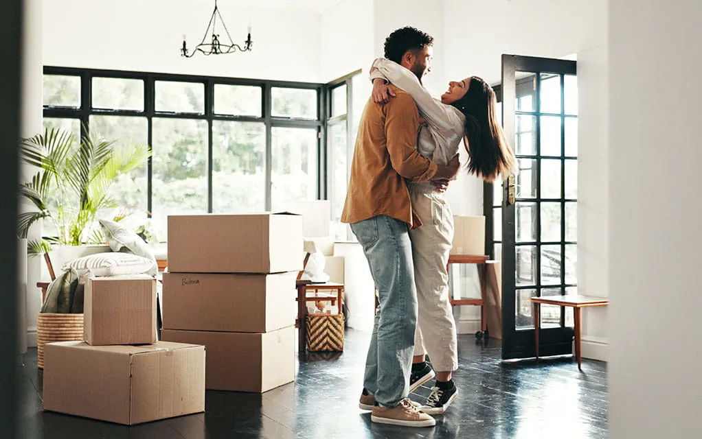 A happy couple holds each other in the foyer of their new home, as moved in boxes full of their belongings sits to the side
