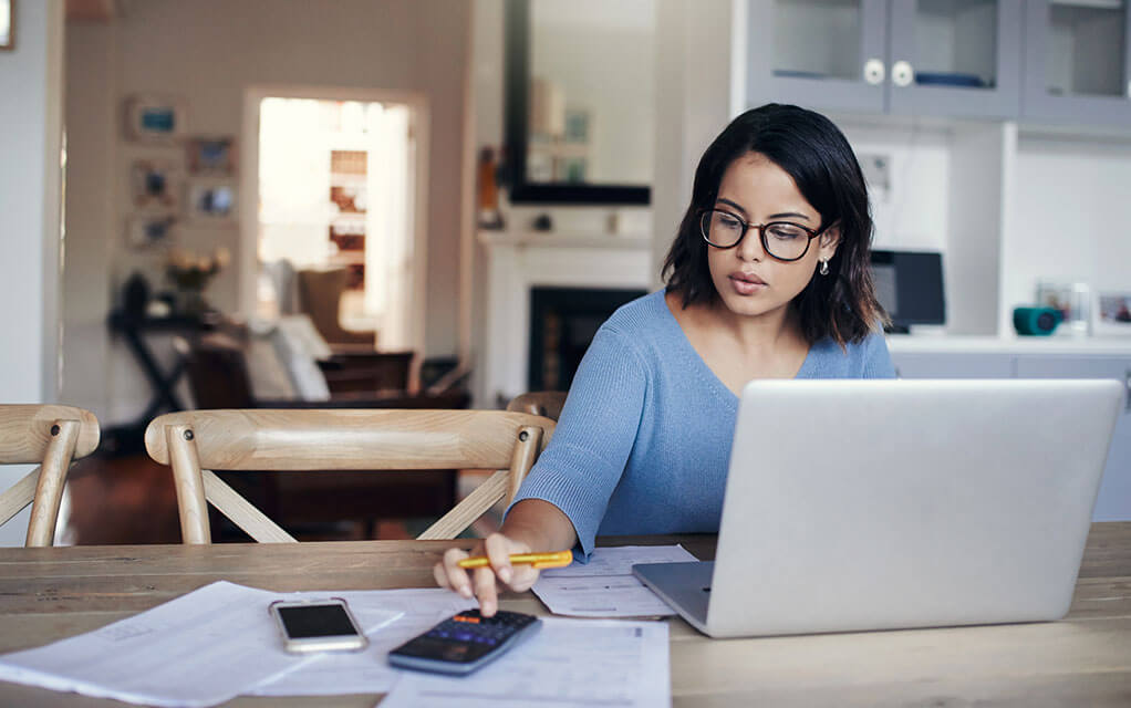A woman sits at her dining room table in her home with a laptop open in front of her as she does calculations on her phone with papers underneath