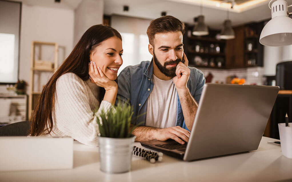 A young couple sits at their kitchen table with a laptop open doing research on something