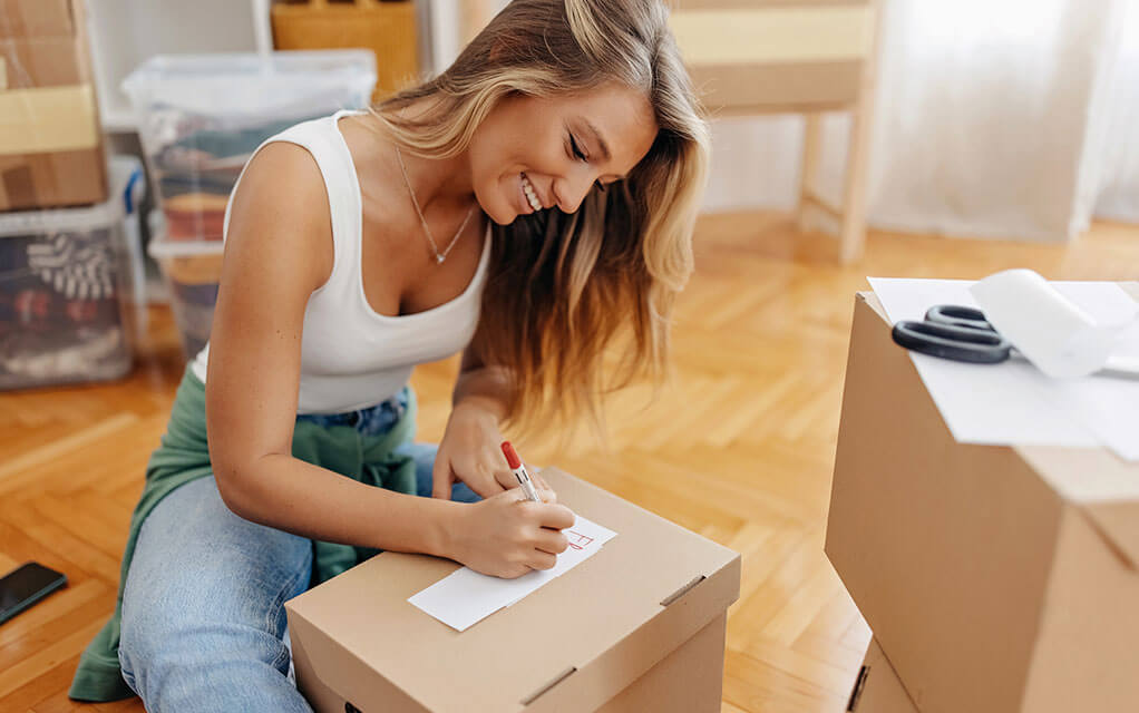 A smiling young woman begins to write down a list of things on a piece of paper on top of a box