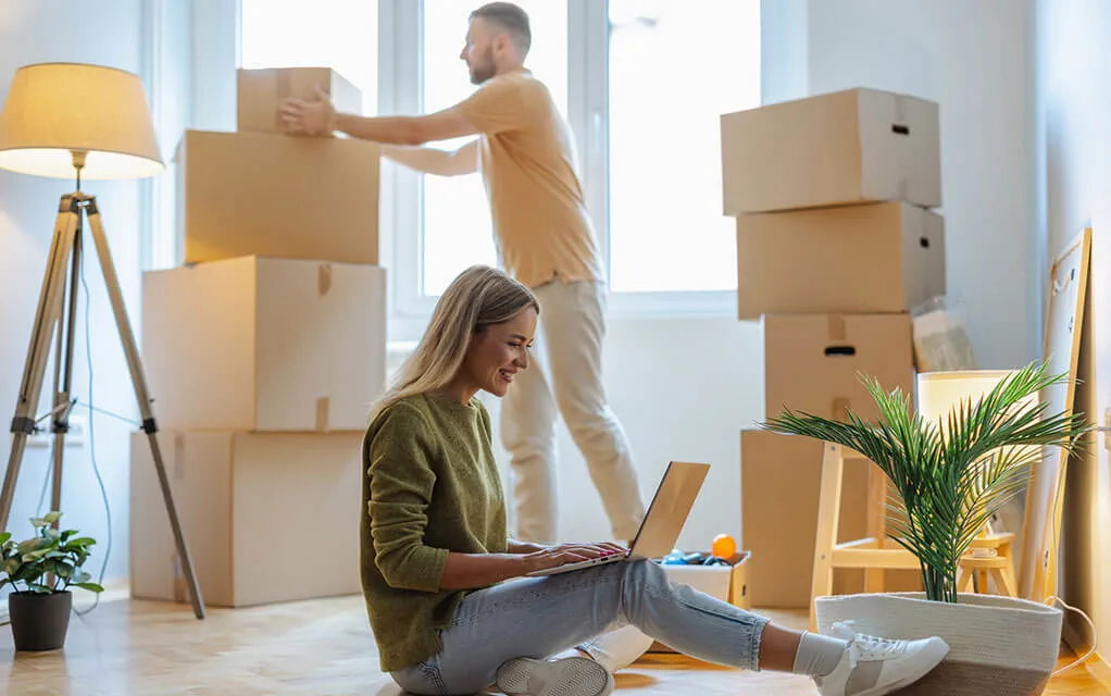 A young couple, a man stacking packing boxes and a woman on the floor with her laptop computer, are in their home's living room getting ready for a move