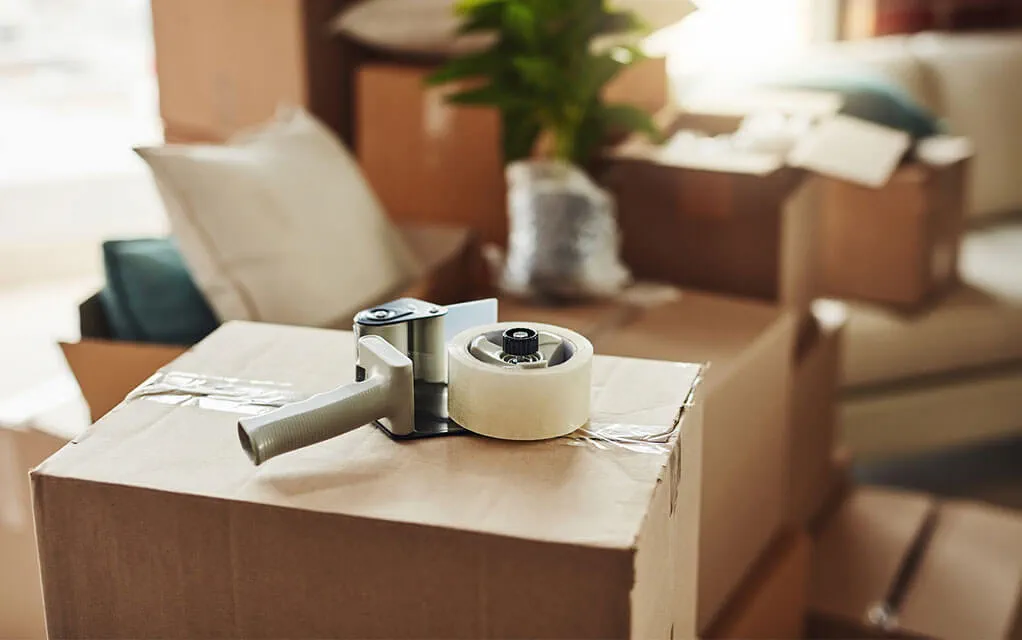 A packing tape gun sits on the top of a closed moving box with more moving boxes and home furniture sitting in the background