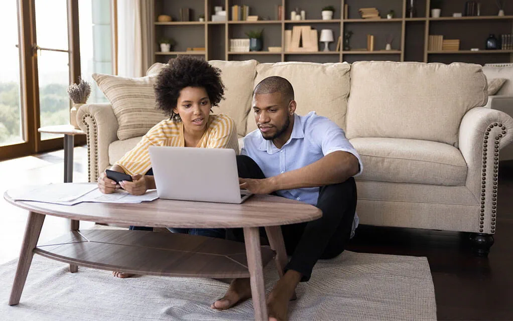 A young couple sits on the floor of their home in between their couch and coffee table with paper work and a laptop on the table