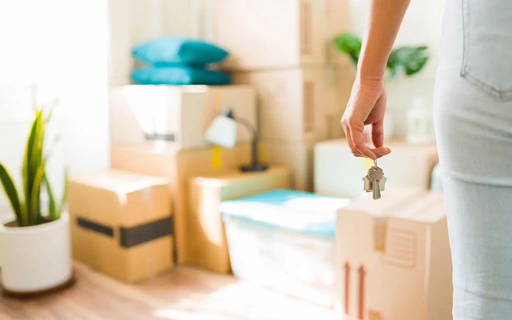 A woman stands in her home's living room as she holds some keys dangling from a keychain as moving boxes sit stacked in the corner of the room with other objects