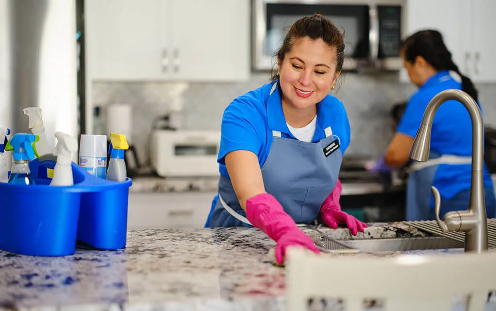 A professional cleaning crew wearing blue uniforms and aprons are cleaning a nice kitchen, as a caddy full of cleaning supplies sits on the kitchen island