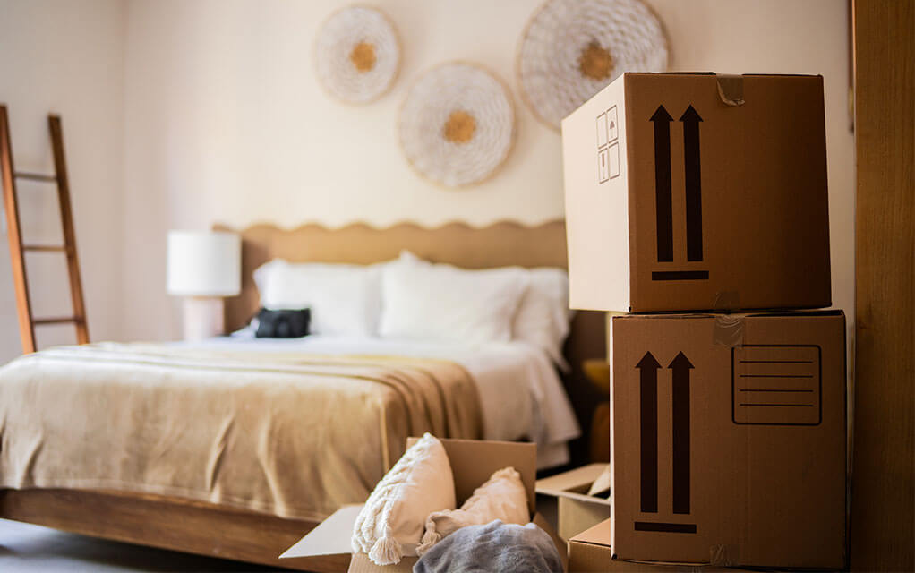 Packed moving boxes sit waiting to be moved in the foreground as a nicely made bed sits in the background in a bedroom