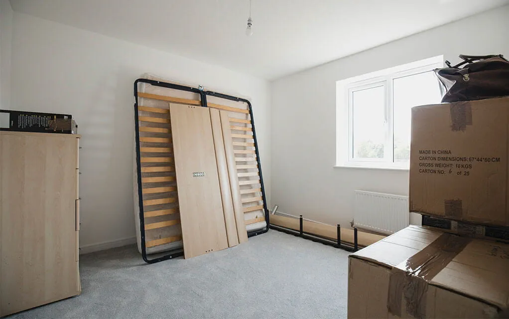 A bed's frame sits against the wall of a bedroom as moving boxes and other furniture wait to be moved out