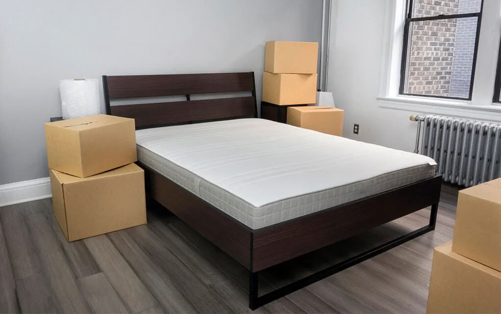 A bed lays in a bedroom with taped up moving boxes stacked up around it