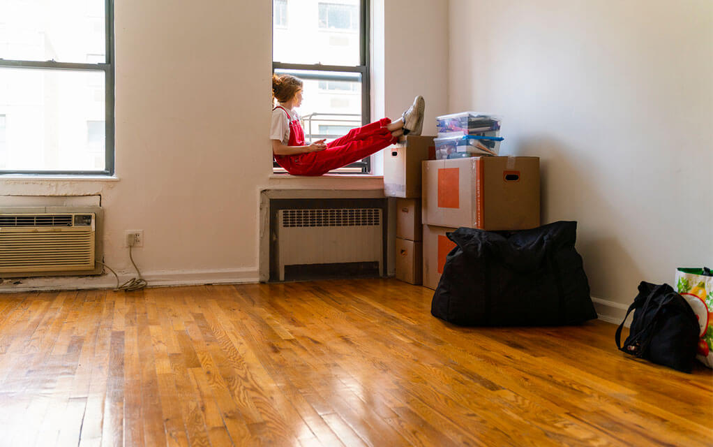 A young woman sits on the windowsill of her small city apartment with her feet propped up on some moving boxes in the corner of the clear room, looking out the window