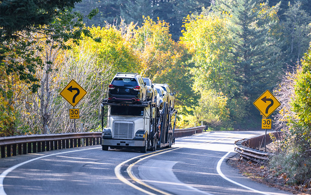 A long-haul open air vehicle carrier truck with multiple vehicles on it drives down a forested two lane highway on a sunny afternoon
