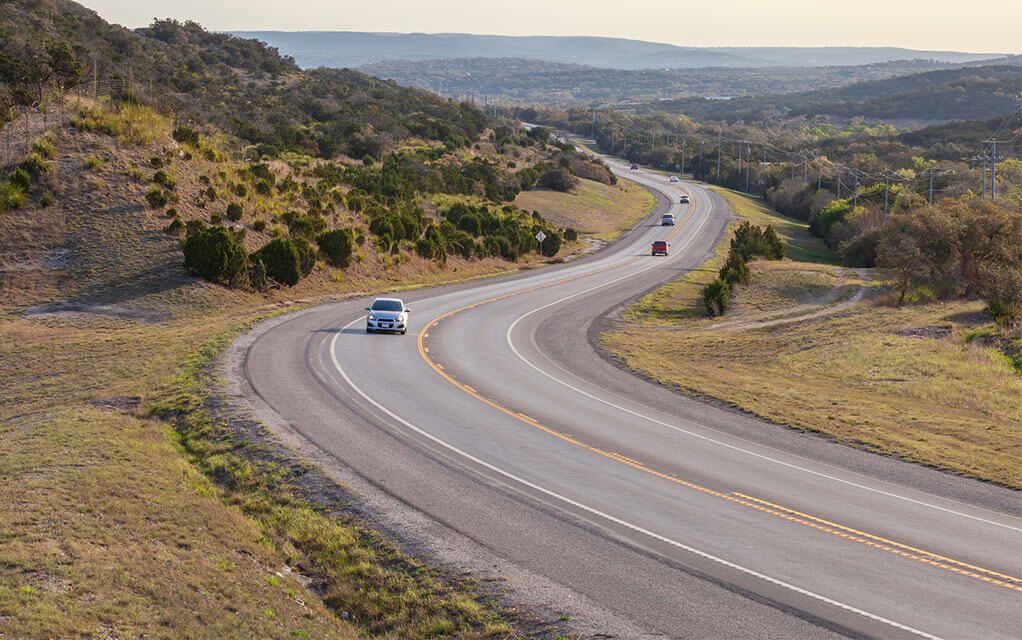 A car travels down a light traffic two-lane highway in the American countryside as multiple vehicles in the background drive the opposite direction at dusk
