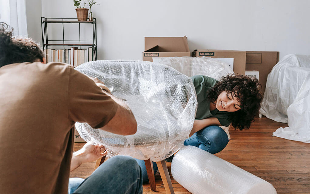 A couple works together to tape bubble wrap around a chair in their living room, as more bubble wrap sits on the floor and more moving boxes and covered furniture lay in the background