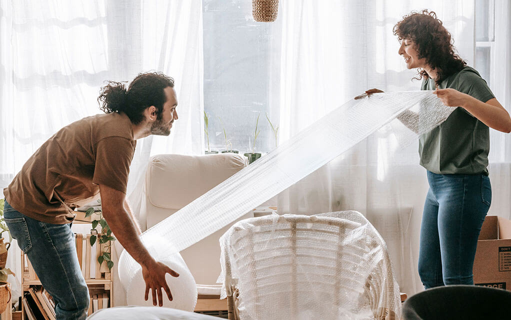 A couple works together to undo some bubble wrap from a large tube of bubble wrap so they can continue to wrap a chair sitting on the floor for their move