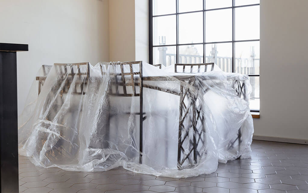 A view of a dining room table and chairs sitting in the open floor plan dining room of a home with a large window behind it as all of the chairs and the table are covered in plastic sheeting