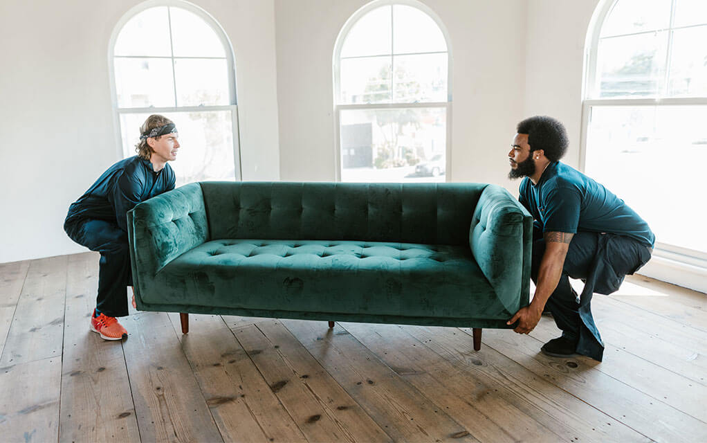 Two male professional laborers are each taking the end of a green velvet couch to pick it up and move it in the middle of a empty living room