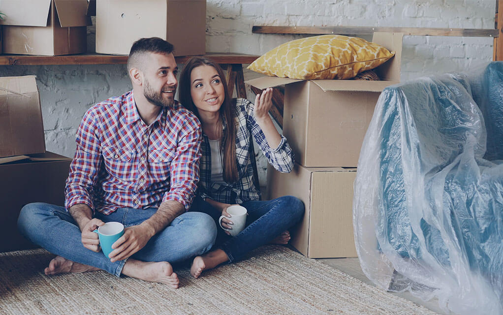A young couple sits on the floor of their home, holding coffee cups and looking around their room as they are surrounded by packed and unpacked moving boxes and a covered blue couch