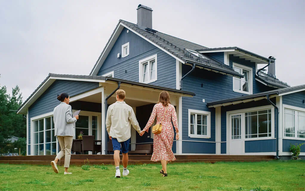 A couple holds hands as they tour a for sale home with a real estate agent, as a blue colored upper middle class home is in the background on a cloudy day