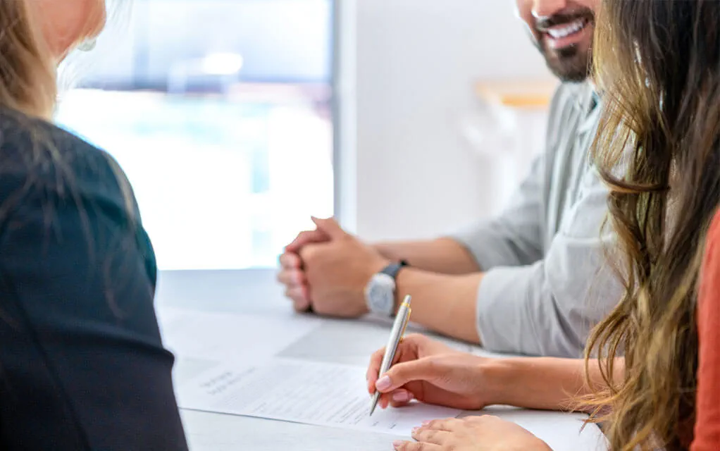 A couple is sitting at a table and is signing documents on a sunny day as a closing agent sits perpendicular to them, speaking to them