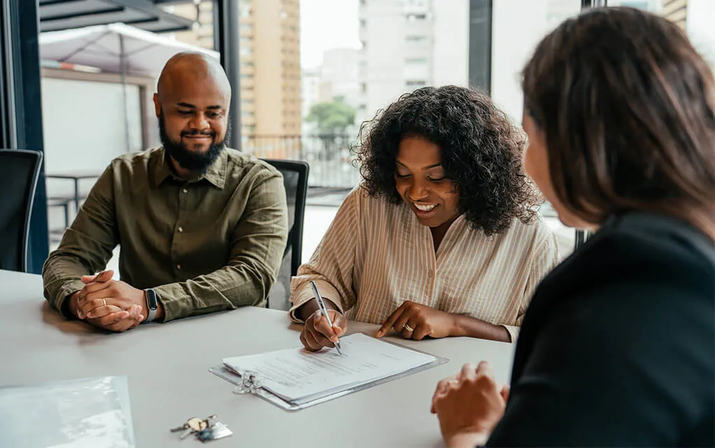 A couple sits and smiles at a conference table and is signing documents as a closing agent speaks with them