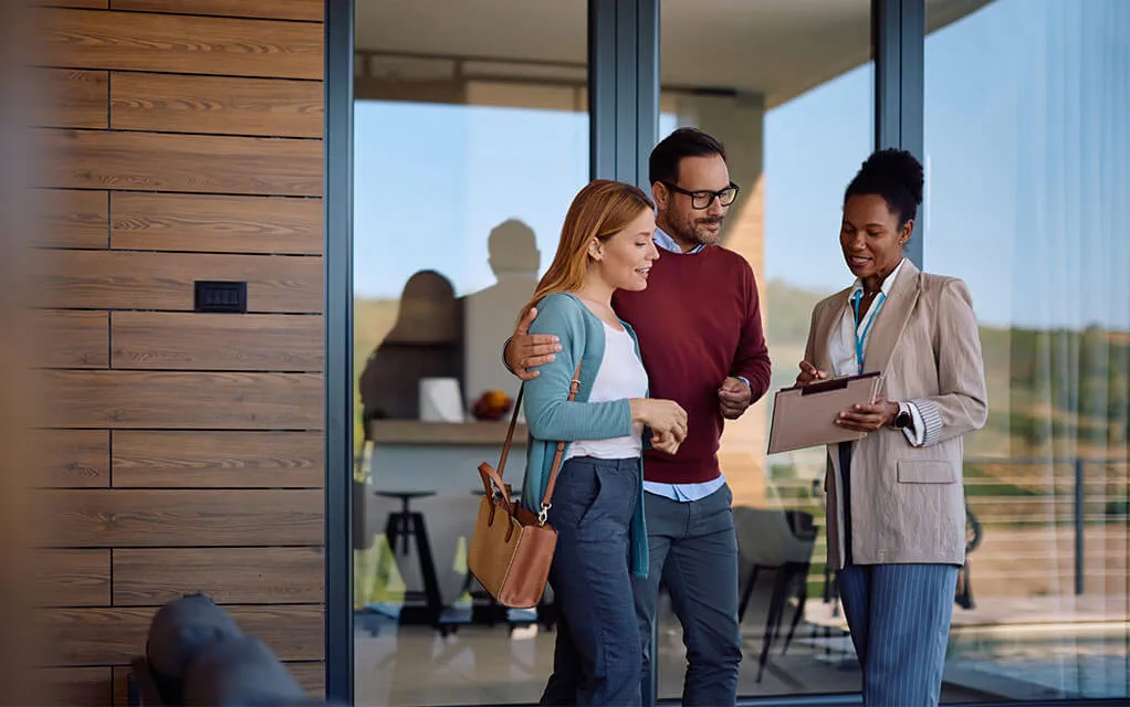 A happy couple talks to a real estate agent who is showing them information on her clipboard as they tour a home they are interested in buying