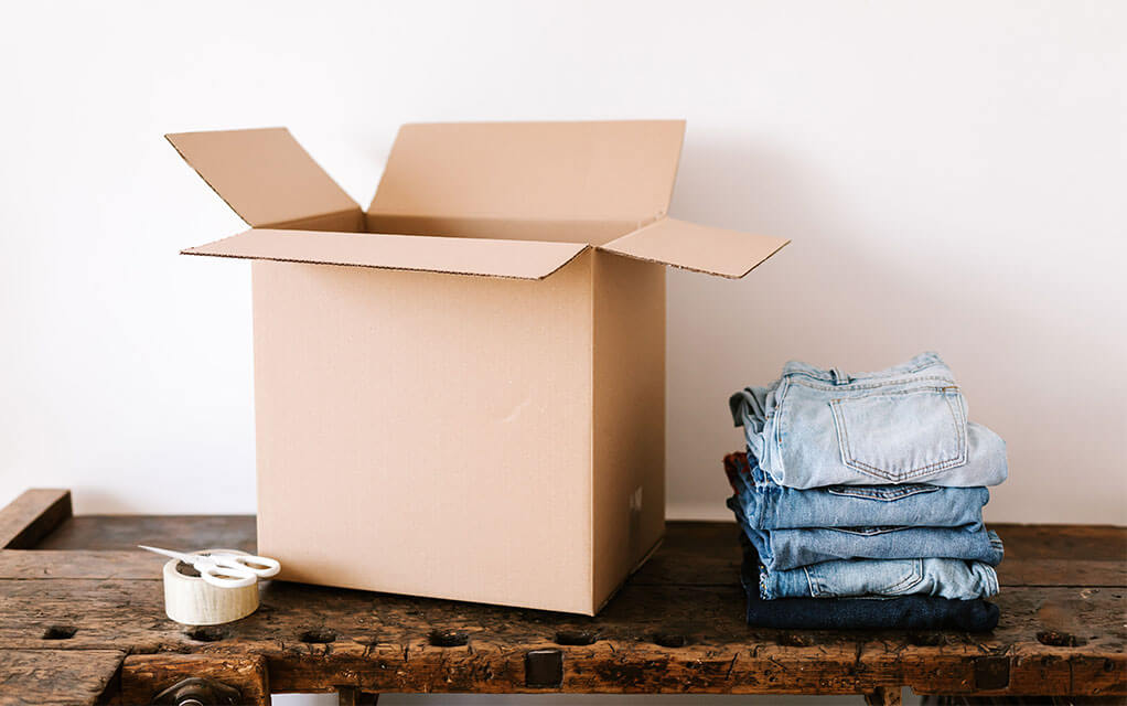 View of an open moving box waiting to be packed with the four pairs of folded jeans sitting on the same table next to it, as a pair of scissors sits on the top of a roll of tape next to the box