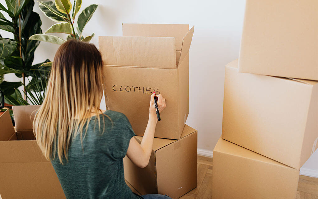 A young woman is packing up stuff for her move and is using a permanent marker to write the word "clothes" on the side of an open moving box next to a three-stack of moving boxes