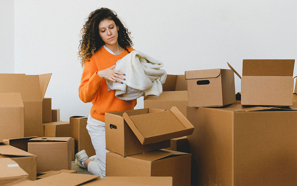 A woman in an orange sweater packs a large sweater into an open moving box, as she is surrounded by a large number of open and closed moving boxes on the floor