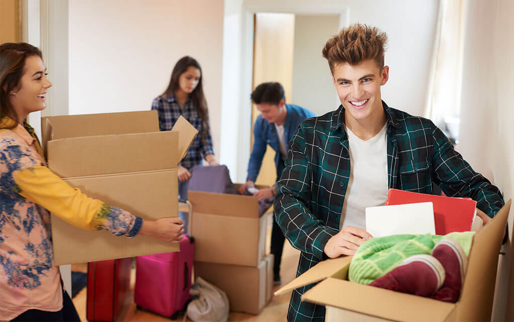 A group of happy, young college students all carry boxes and unpack them in a dorm suite as opened boxes lay scattered on the floor of the brightly lit room