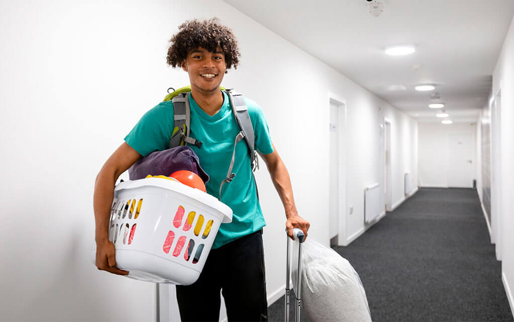 A young college student carrying a laundry basket with random items and carrying a suitcase with a laundry bag and a backpack walks and smiles in the hallway of his dormitory
