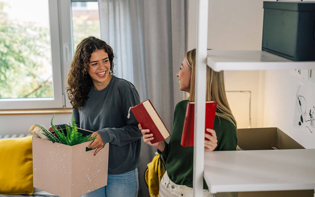 Two young women laugh as one places books on a shelf and the other carries a box with a plant and other items inside it as they unpack their shared dorm room