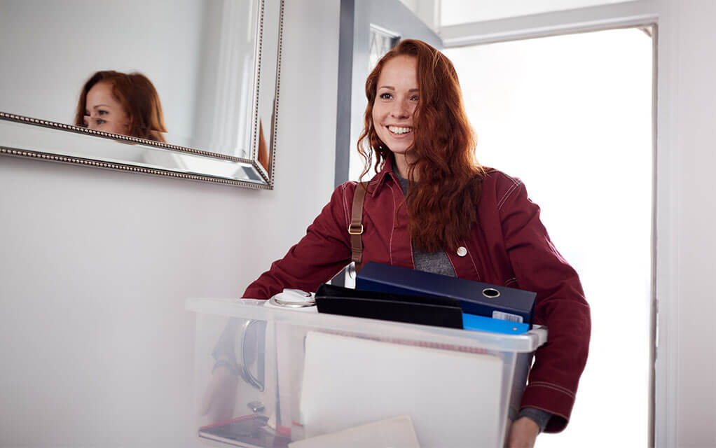 A young woman carrying a large plastic bin filled with her belongings from college smiles as she walks her stuff into her home