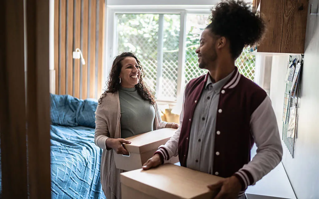 A young college student and his mother smile at each other as they carry packed boxes out of his dorm room.