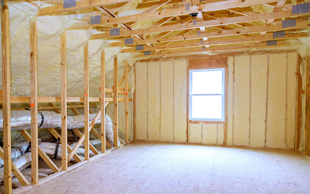 View of an empty attic space with foam insulation sprayed between the wall studs and the ceiling rafters and joists as sunlight streams through the window