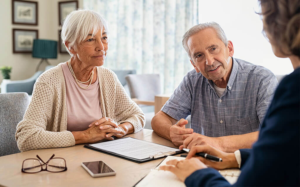 An elderly couple speaks with a lawyer about a legal matter, as documents sit on a clipboard and a phone and pair of glasses sits near them