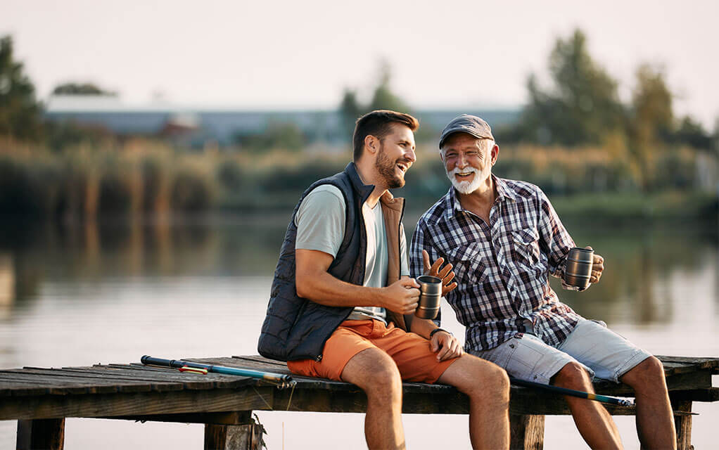 An adult man and his elderly father sit on a pond doc on a clear evening as they share a drink and laugh as they talk with each other