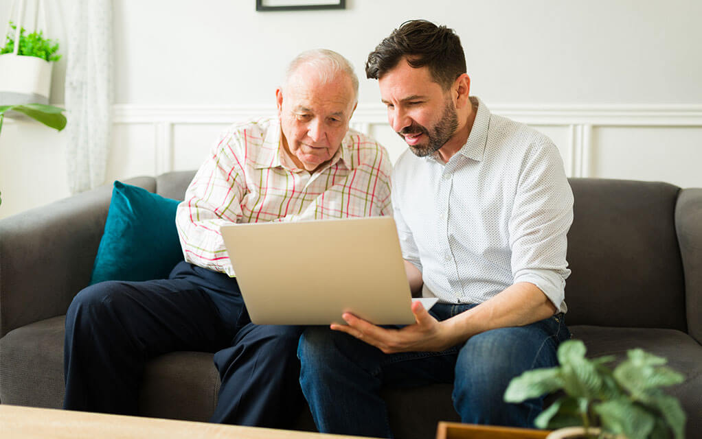 An adult man and his elderly father look at something on a laptop as they sit on a couch in a living room on a bright day