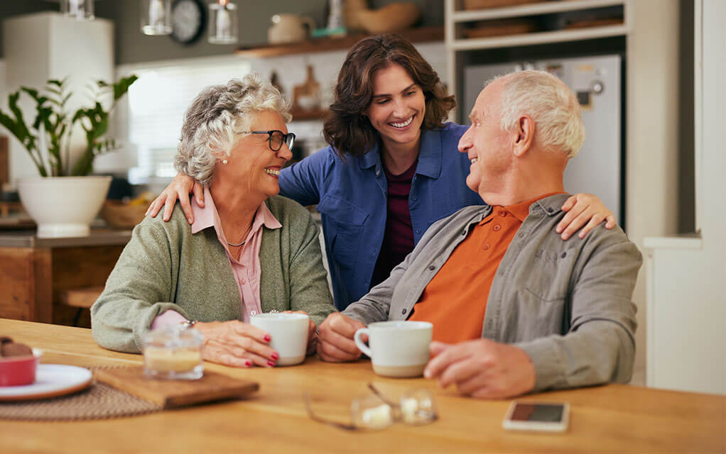 An adult woman puts her arms around her elderly parents sitting at a big kitchen table as they drink coffee